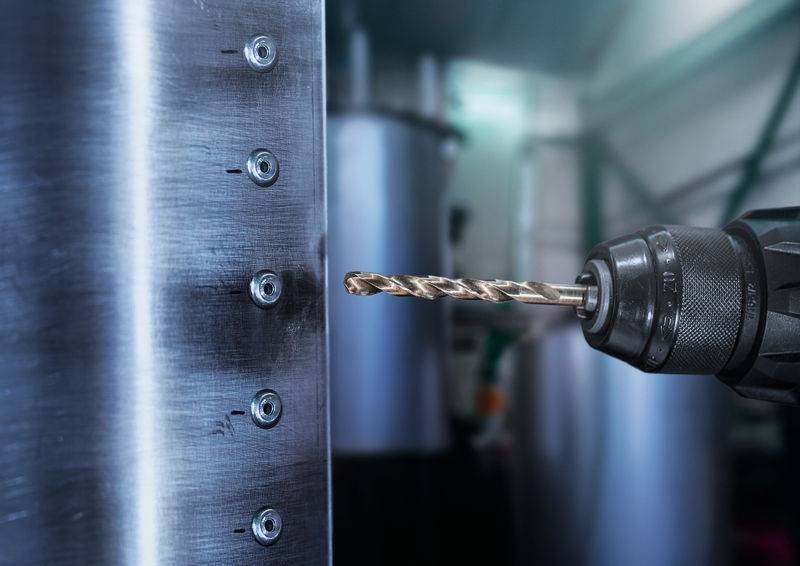 Eine Nahaufnahme eines Bohrers, der Löcher in ein Metallblech bohrt, mit unscharfen Industriemaschinen im Hintergrund.