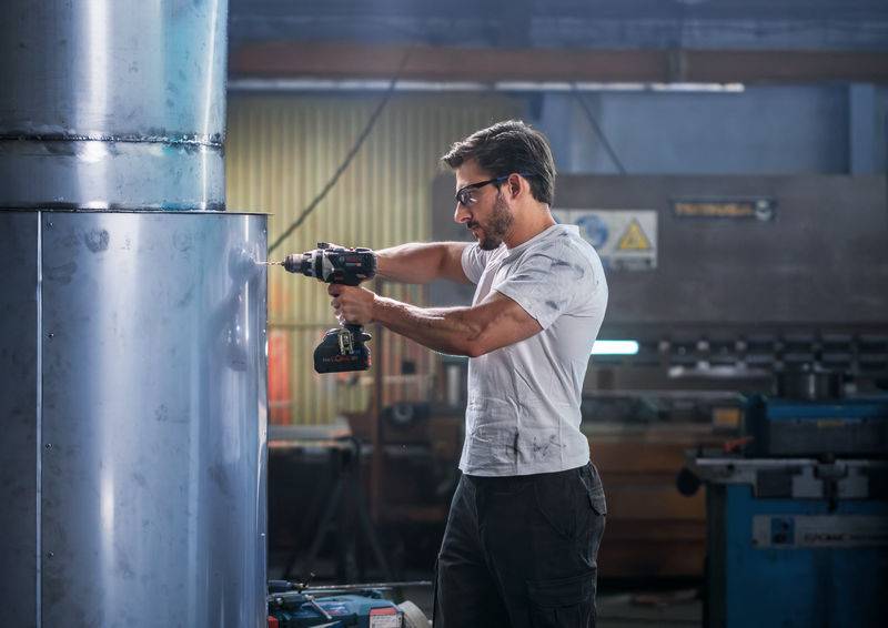Ein Mann in einer Werkstatt benutzt einen Elektroschrauber an einem großen Metallzylinder, umgeben von Industrieausrüstung, was ein Gefühl von Handwerkskunst vermittelt.
