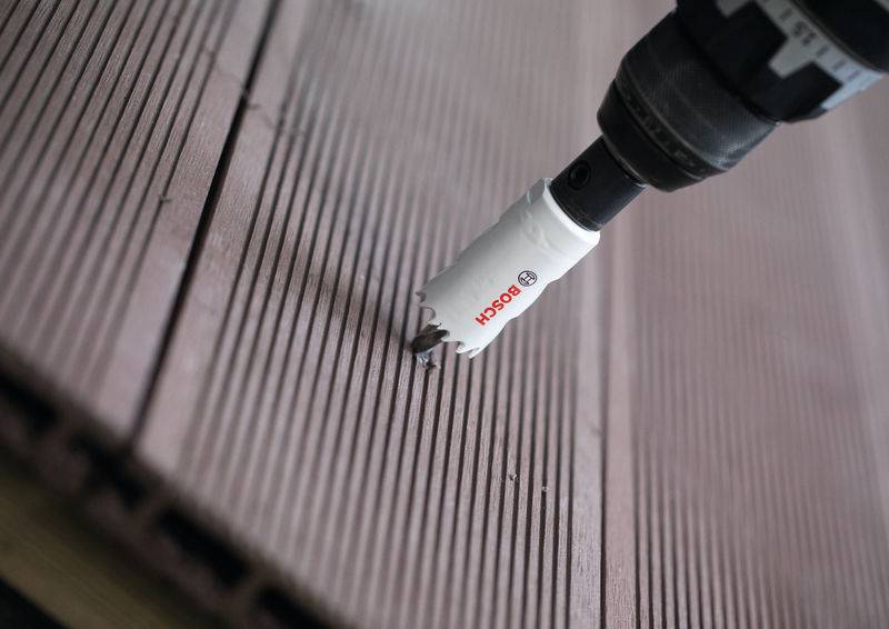 Ein Elektrobohrer mit einem Bosch-Lochsägen-Aufsatz ist positioniert, um ein Loch in eine braune, gerillte Holzplanke eines Decks zu schneiden.