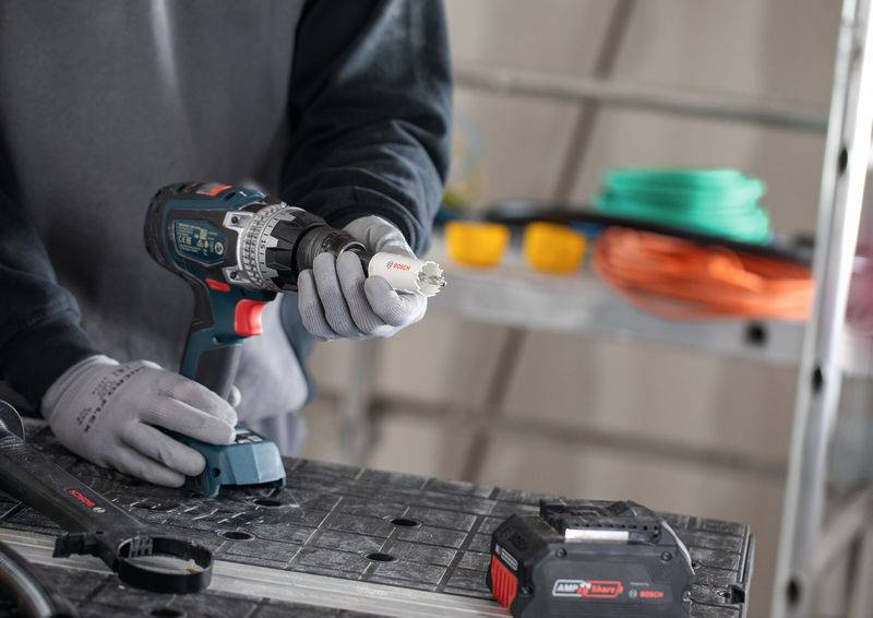 Eine Person mit Handschuhen befestigt ein Werkzeug an einem Elektrobohrergerät. Im Hintergrund auf einer Werkbank sind aufgerollte Kabel und ein Akku sichtbar.