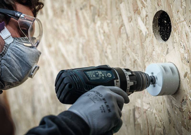 Ein Arbeiter in Schutzausrüstung bohrt mit einer Elektrobohrmaschine mit Lochsägenaufsatz ein Loch in ein Holzbrett.
