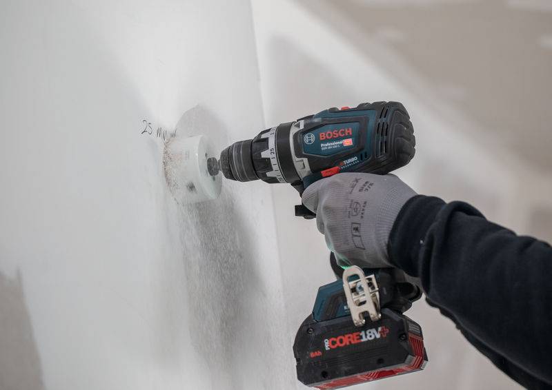 Eine Person nutzt eine Bosch CORE18V-Bohrmaschine, um in eine weiße Wand zu bohren, trägt dabei Schutzhandschuhe und zeigt damit die Verwendung von Elektrowerkzeugen im Bauwesen.