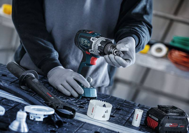 Eine Person mit Handschuhen hält einen kabellosen Elektroschrauber auf einer Werkbank mit verschiedenen Werkzeugen und Bohrern, die um sie herum angeordnet sind.