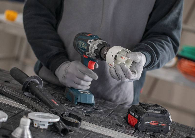 Eine Person, die einen kabellosen Bohrer verwendet, um einen Lochsägenaufsatz anzubringen und sich auf eine Bohraufgabe vorbereitet. Werkbank und Bohrer-Akku sind in der Nähe sichtbar.