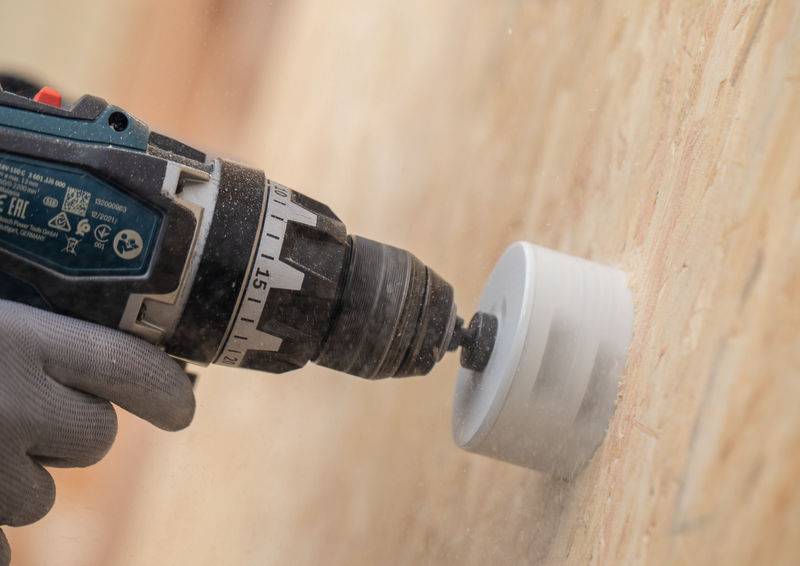 Eine Person benutzt einen Elektroschrauber mit einer Lochsäge-Aufsatzfütterung, um ein Loch in eine hellgefärbte Wand zu bohren und trägt dabei einen grauen Handschuh zum Schutz.