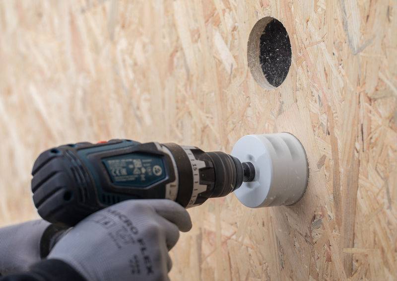 Eine Person, die Handschuhe trägt, verwendet einen Elektroschrauber mit einer Lochsäge, um eine kreisförmige Öffnung in einem Sperrholzbrett zu schaffen.