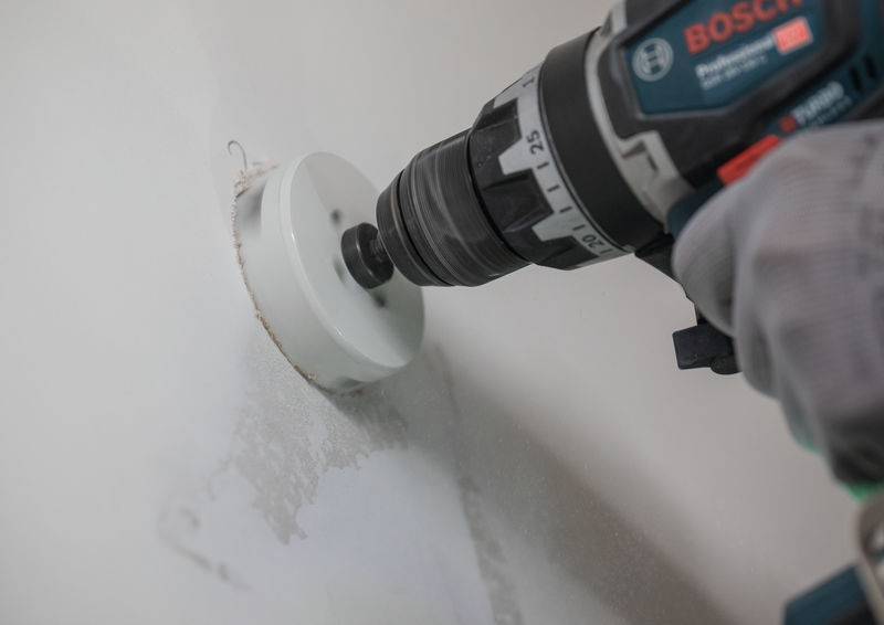 Ein Bohrer mit einer Lochsägen-Aufsatzvorrichtung erzeugt ein kreisförmiges Loch in einer Wand. Die Person, die den Bohrer bedient, trägt Schutzhandschuhe.