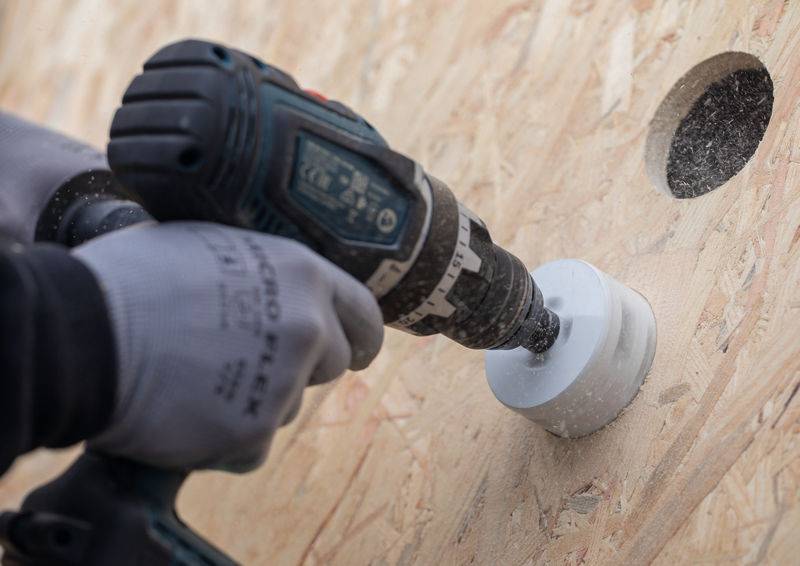 Eine Person mit Handschuhen bohrt mit einem Elektrowerkzeug ein kreisrundes Loch in eine Holzplatte. Staub ist um das neu geschnittene Loch sichtbar.