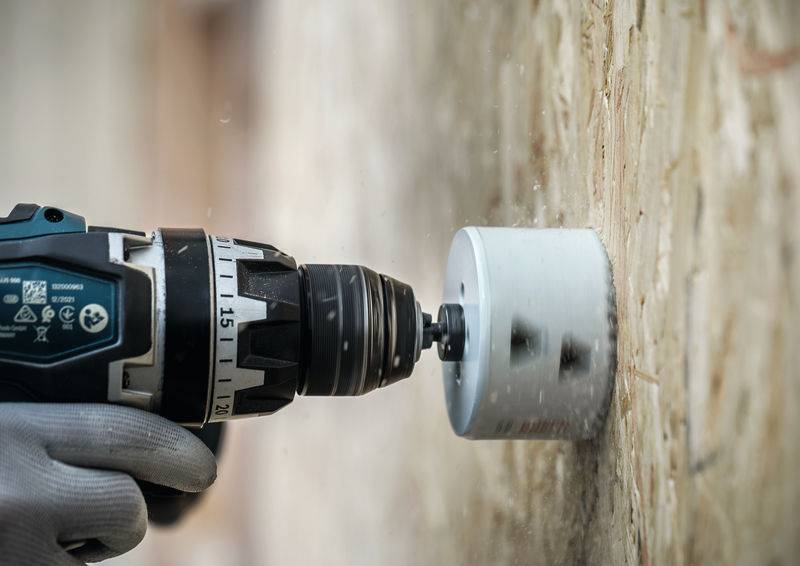 Eine Person trägt Handschuhe und benutzt eine Elektrobohrmaschine mit einem Lochsägen-Aufsatz, um präzise in eine Holzoberfläche zu bohren.