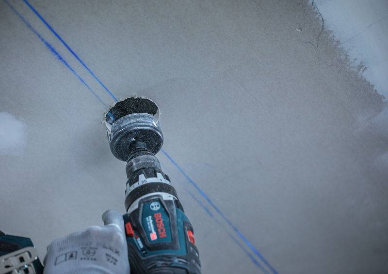 Eine Person benutzt einen Elektrobohrer mit einem Lochsägen-Aufsatz, um in eine Trockenbaudecke zu schneiden, die mit einer blauen Richtlinie für Präzision markiert ist.