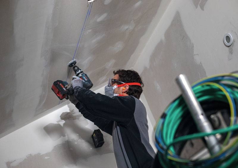 Eine Person, die Schutzausrüstung trägt, benutzt ein Elektrowerkzeug an einer Trockenbaudecke in einer Baustelle. Aufgerollte Kabel sind im Vordergrund sichtbar.