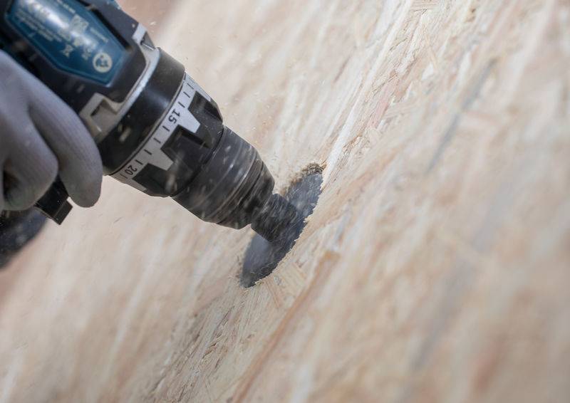 Ein Bohrer wird verwendet, um ein kreisförmiges Loch in einer Holzoberfläche zu schaffen. Die Person trägt Handschuhe und betont damit die Sicherheit bei der Arbeit.