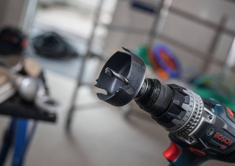 Ein Elektrobohrer mit einer Lochsäge-Aufsatzvorrichtung steht im Fokus vor einem Baustellen-Hintergrund, was auf aktive Verwendung bei einem Bau- oder Reparaturprojekt hindeutet.