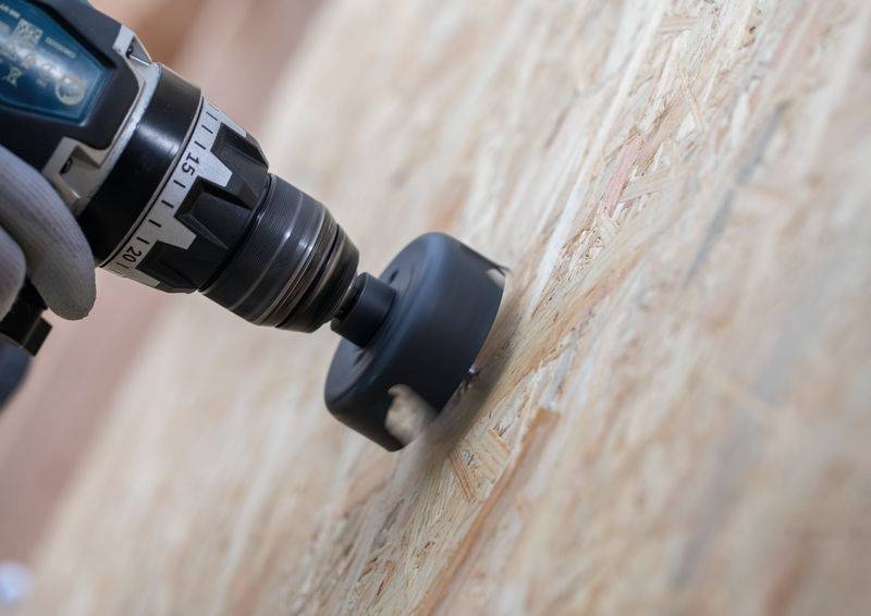 Nahaufnahme einer Elektrobohrmaschine mit einem Lochsägenaufsatz, die durch ein Holzbrett schneidet und Schreiner- oder DIY-Arbeiten demonstriert.