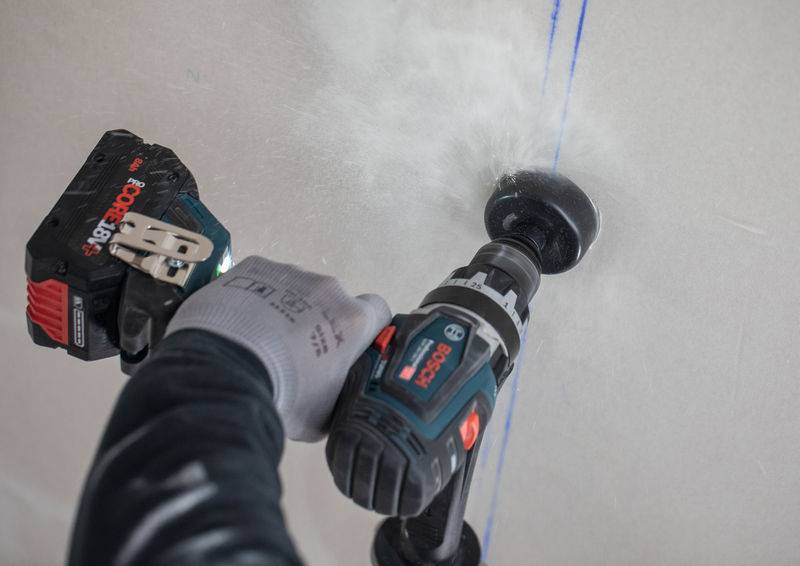 Eine Person benutzt einen kabellosen Elektroschrauber, um ein Loch in einer Gipsplatte zu bohren, wobei Staub sichtbar verteilt wird. Die Hand ist durch einen Handschuh geschützt.