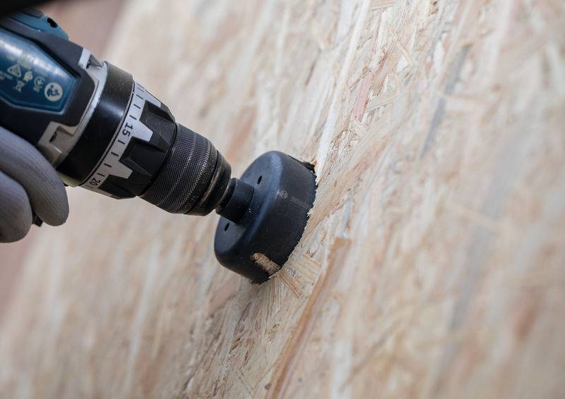 Eine behandschuhte Hand nutzt einen Elektroschrauber mit einem Lochsägenaufsatz, um ein kreisförmiges Loch in eine Platte aus Oriented Strand Board (OSB) zu schneiden.
