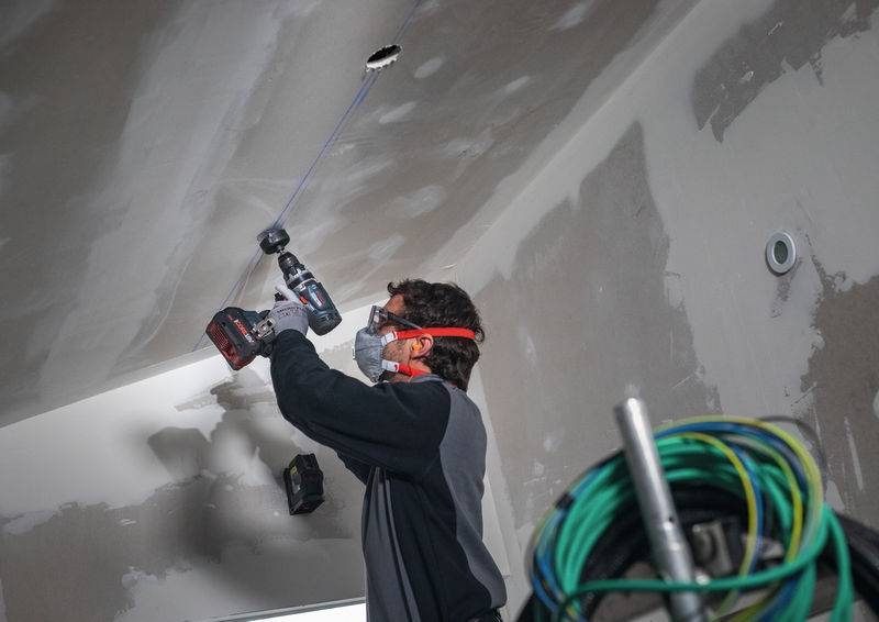 Eine Person mit Schutzbrille und Maske bohrt in einem Konstruktionsumfeld in eine Decke, mit aufgerollten Kabeln in der Nähe sichtbar.