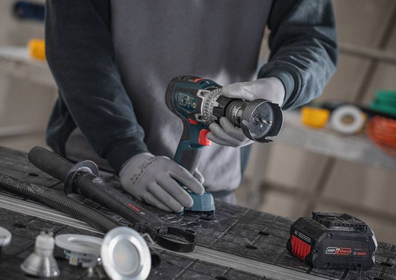 Eine Person mit Handschuhen benutzt einen kabellosen Bohrer auf einer Werkbank, die von verschiedenen Werkzeugen und Komponenten umgeben ist, was auf eine Konstruktions- oder Reparaturaufgabe hindeutet.