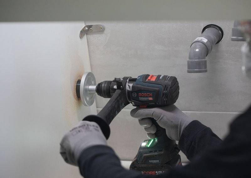 Eine Person bohrt mit einem schwarz-roten Elektrobohrer ein Loch in eine weiße Wand. Im Hintergrund sind Rohre sichtbar.