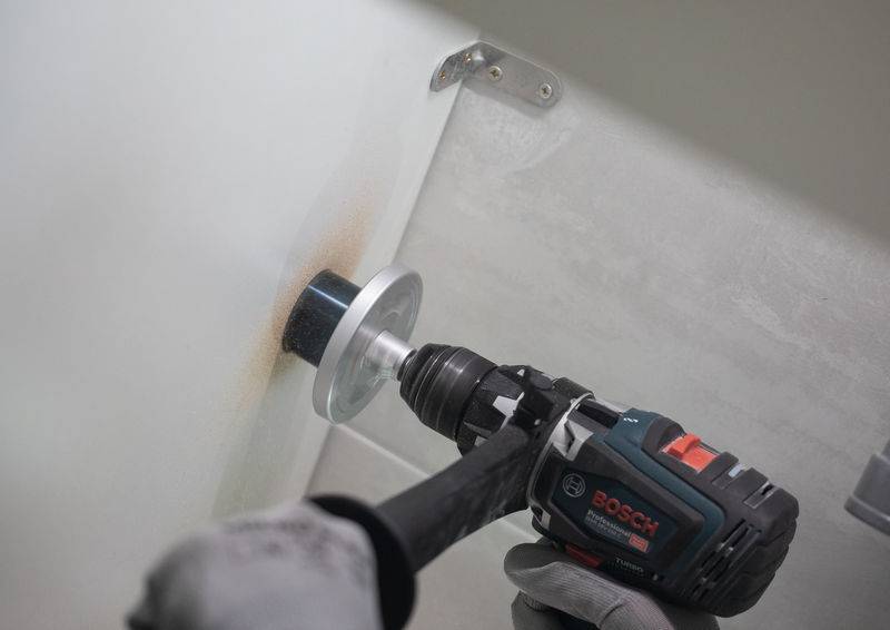 Eine Person, die einen Bosch-Elektrobohrer mit einer Lochsägen-Aufsatzbestückung verwendet, um ein Loch in eine weiße Wand zu bohren. Der Bohrer wird fest gegen die Oberfläche gepresst.