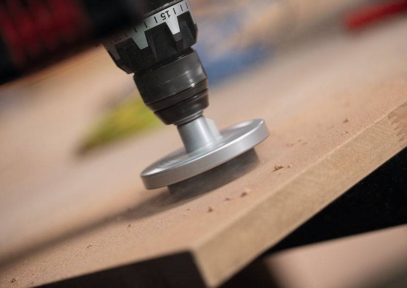 Ein Bohrer mit einer Schleifaufsatz glättet die Kante eines Holzbretts und erzeugt dabei Holzstaub.