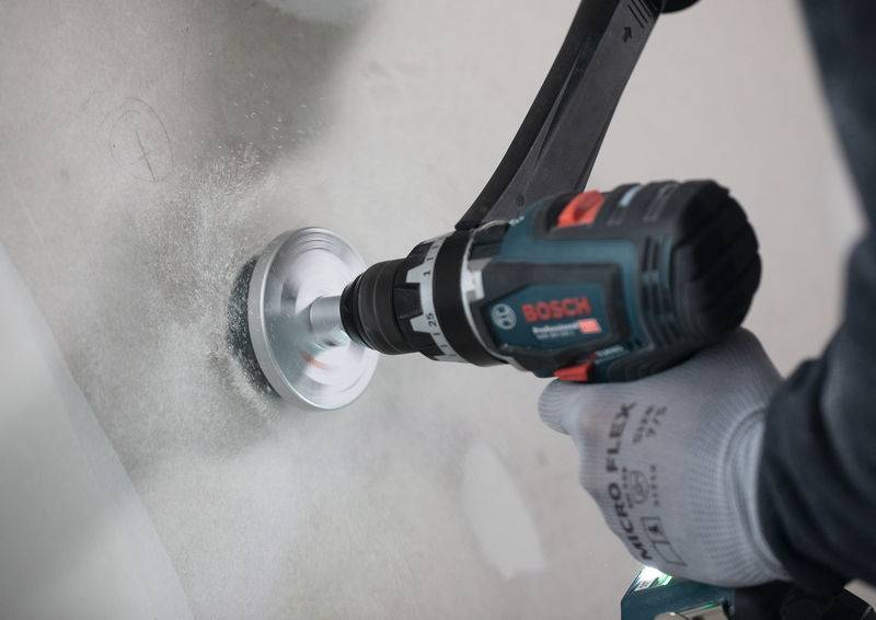 Eine Person mit Handschuhen benutzt eine Bosch Elektrobohrmaschine, um ein Loch in eine Wand zu bohren, wobei während des Prozesses Staub entsteht.