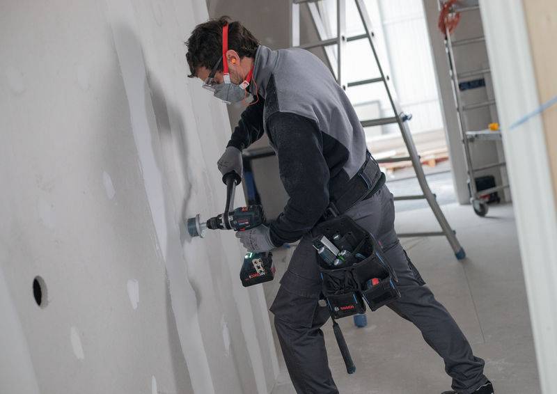 Ein Arbeiter in Schutzausrüstung bohrt mit einem tragbaren Elektrobohrer ein Loch in eine Gipskartonplatte in einer Baustelle.