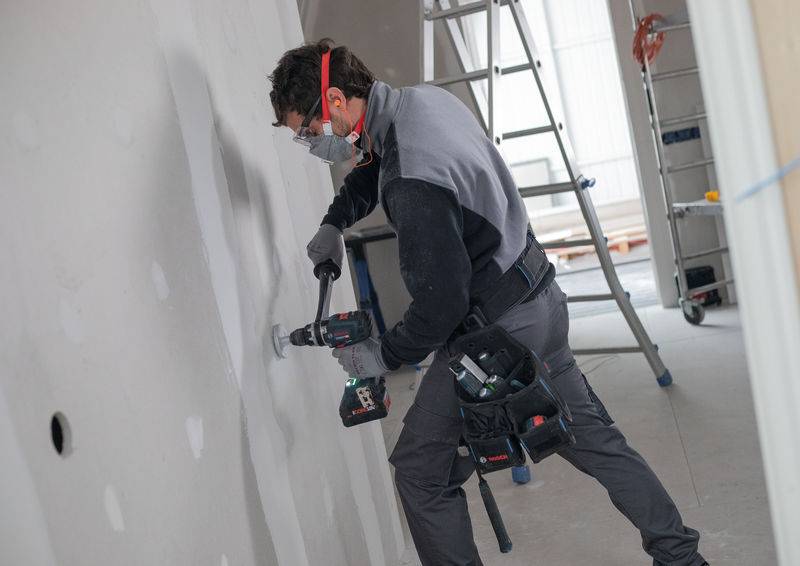 Bauarbeiter, der einen Bohrer an einer Gipskartonwand in einem Raum unter Renovierung verwendet, trägt Schutzausrüstung und ist von Leitern und Werkzeugen umgeben.