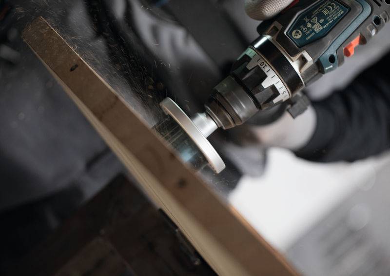 Eine Person benutzt eine elektrische Bohrmaschine mit einer kreisförmigen Bürstenaufsatz auf einer Holzoberfläche und erzeugt dabei eine Staubwolke aus Sägespänen in einer Werkstattatmosphäre.