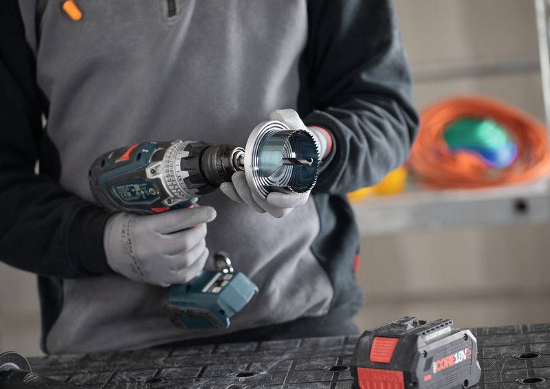 Eine Person mit Handschuhen benutzt einen Elektroschrauber, um ein Kreissägeblatt anzubringen. Im Hintergrund ist ein orangefarbenes Verlängerungskabel sichtbar.