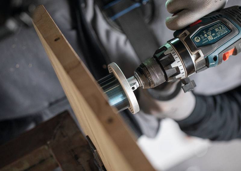 Eine Person mit Handschuhen benutzt einen Elektroschrauber, um ein Loch in ein Holzbrett zu bohren. Der Fokus liegt auf der Wechselwirkung zwischen dem Bohrmeißel und dem Holz.