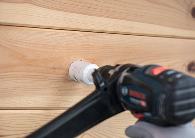 Eine behandschuhte Hand benutzt einen Bosch-Bohrer, um ein Loch in ein helleres Holzbrett zu bohren. Das Brett zeigt sichtbare natürliche Maserungsmuster.