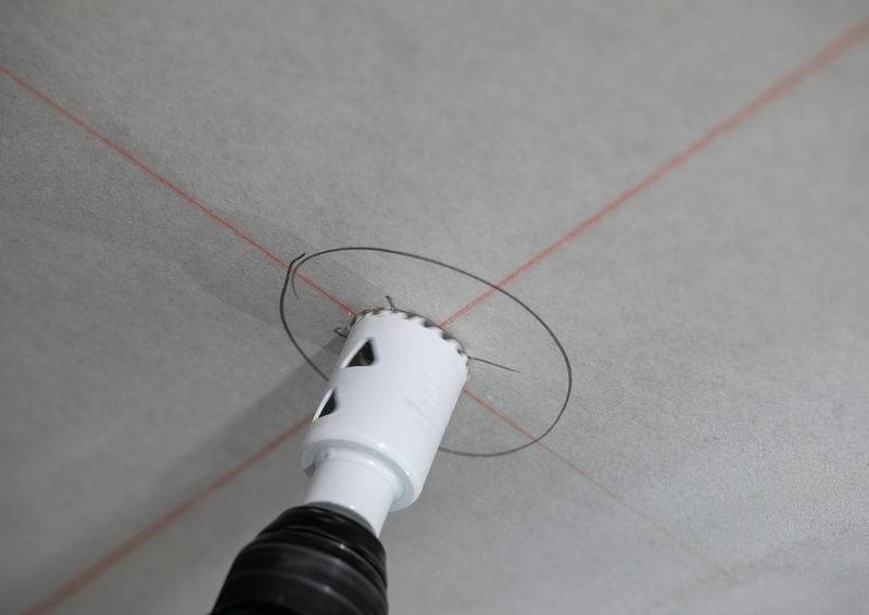 Ein Bohrer mit einer Lochsäge-Aufsatz erzeugt einen kreisförmigen Schnitt in einer Decke, ausgerichtet an roten Laserführungslinien für präzise Positionierung.