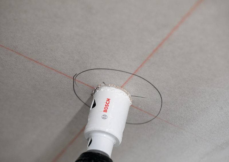Ein Bohrgerät mit einer Lochsäge schneidet in eine Decke ein, die mit sich kreuzenden roten Laserlinien zur Präzision gekennzeichnet ist.