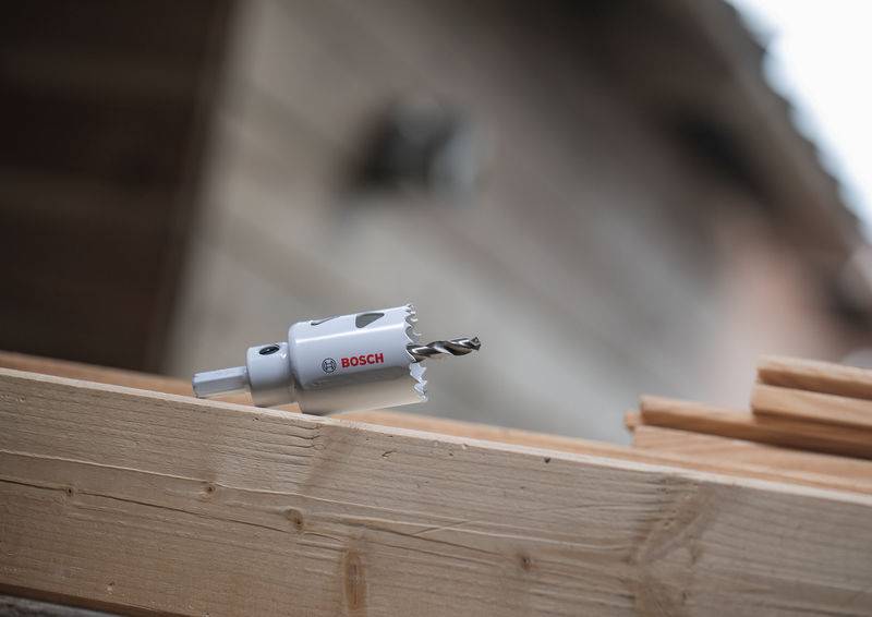 Ein Lochsägen-Bohraufsatz ist an einem Elektrowerkzeug befestigt und ruht auf einem Holzbalken im Freien vor einem verschwommenen Gebäudehintergrund.