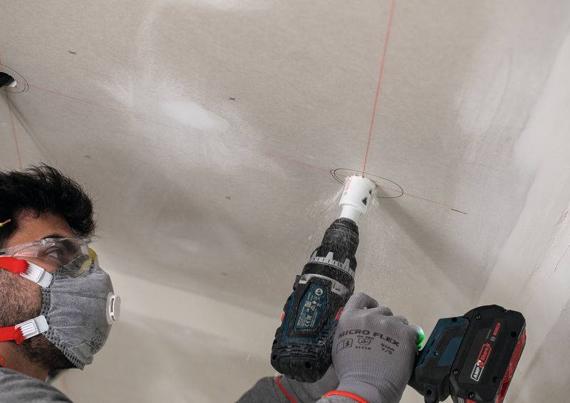 Eine Person trägt eine Schutzbrille und eine Staubmaske und verwendet einen Bohrer, um ein kreisförmiges Loch in eine Decke zu schneiden, wobei sie sich an roten Laserführungslinien ausrichtet.