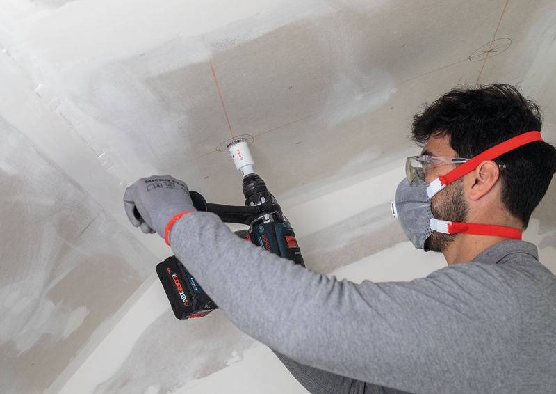 Eine Person, die eine Maske und eine Schutzbrille trägt, benutzt einen Elektroschrauber, um an einer Decke zu arbeiten. Die Decke scheint sich im Bau oder in der Renovierung zu befinden.