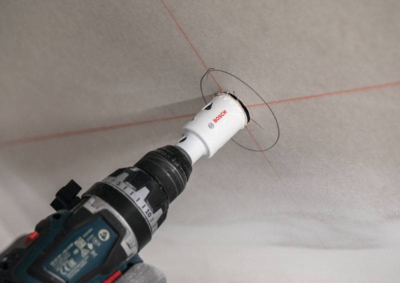 Ein Elektroschrauber mit einer Lochsäge-Aufsatzklinge schneidet in eine weiße Decke, wobei rote Führungslinien sichtbar sind.
