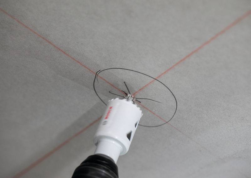 Ein Bohrer mit Kreissägen-Aufsatz erstellt einen präzisen Kreis auf einer grauen Oberfläche, die mit roten Markierungslinien für genaue Positionierung versehen ist.