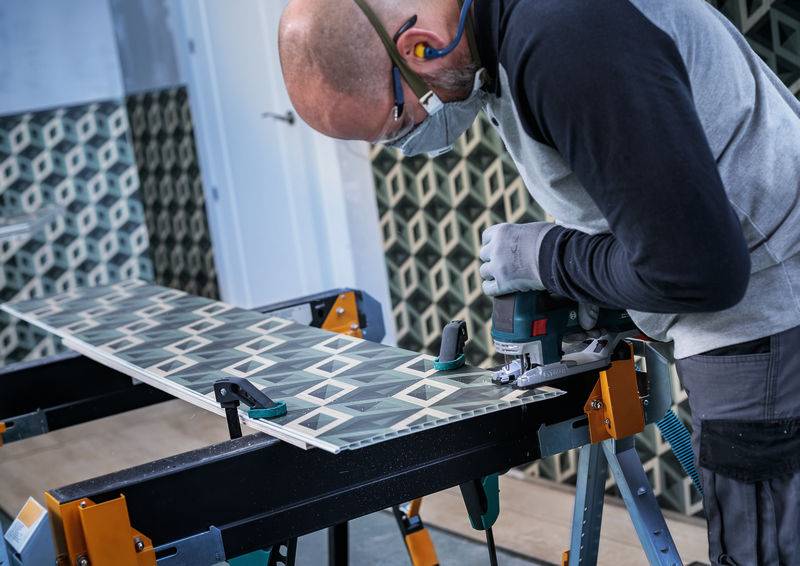 Eine Person in Schutzausrüstung nutzt ein Elektrowerkzeug, um eine gemusterte Fliese auf einer Werkbank in einem gefliesten Raum zu schneiden, was auf Renovierungsarbeiten hindeutet.