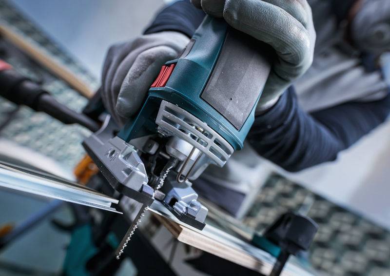 Eine Person trägt Handschuhe und benutzt eine elektrische Stichsäge, um ein Metall- oder Holzblatt zu schneiden. Die Szene konzentriert sich auf die Schneidaktion.