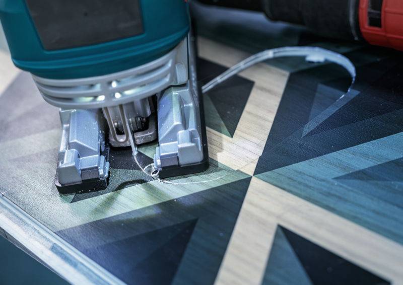 Eine Elektrostichsäge schneidet ein geometrisches, dunkles Holzmuster. Die Nahaufnahme zeigt Sägespäne und die Präzision des gezackten Blattes auf der Oberfläche.