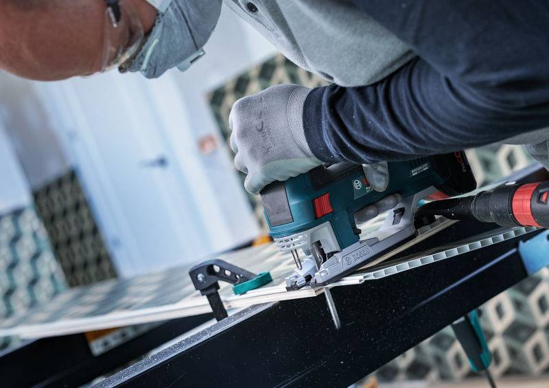 Eine Person benutzt ein Elektrosägewerkzeug auf einem Materialblatt, trägt Schutzhandschuhe und eine Maske, während Spannzwingen das Material auf einem Tisch sichern.
