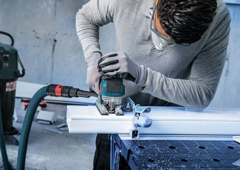 Eine Person, die mit einer Stichsäge ein Stück weißes Material auf einer Werkbank schneidet, trägt dabei eine Schutzbrille und Handschuhe in einer Werkstattumgebung.