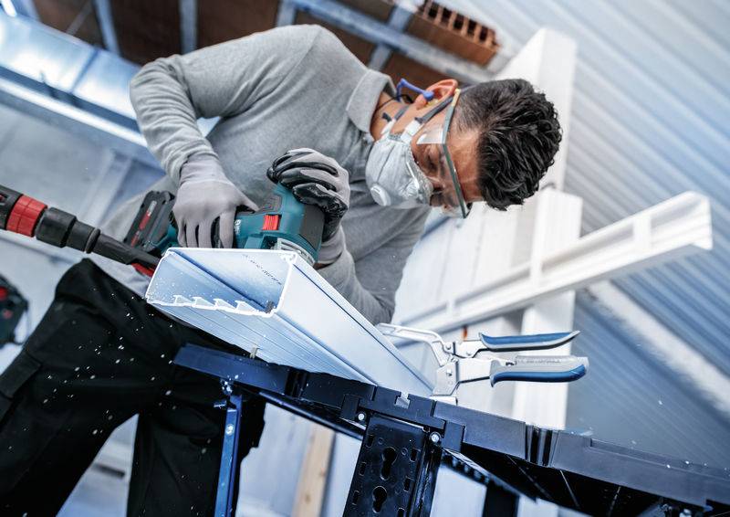 Eine Person in Schutzausrüstung, einschließlich Schutzbrille und Maske, benutzt ein Elektrowerkzeug, um Metall- oder Kunststoffplatten in einer Werkstatt zu schneiden.