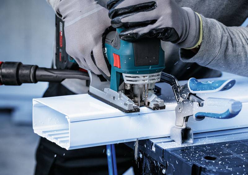 Eine Person benutzt eine Elektrosäge, um ein weißes Material zu schneiden, das mit einer Klemme gesichert ist, und trägt Sicherheitshandschuhe.