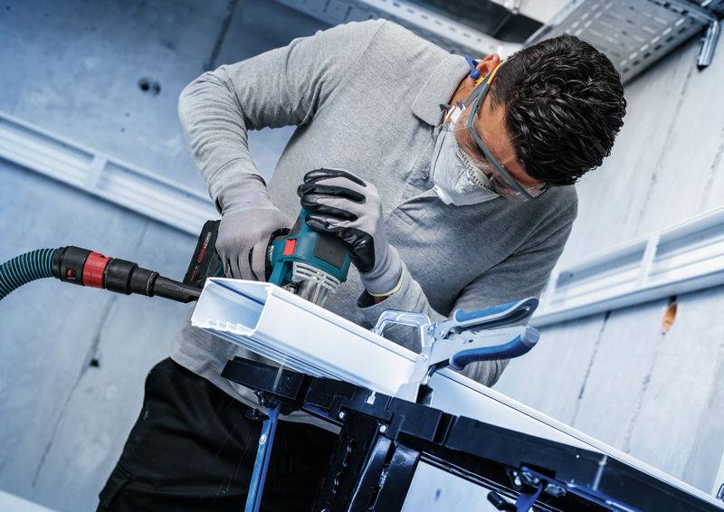 Eine Person, die eine Schutzbrille und eine Maske trägt, verwendet einen Elektroschrauber auf einem Materialstück und konzentriert sich dabei in einer Werkstattumgebung.