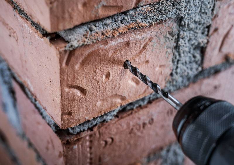 Nahaufnahme eines Bohrers, der ein Loch in eine rote Backsteinmauer bohrt, umgeben von grauem Mörtel, zeigt Bau- oder DIY-Aktivität.
