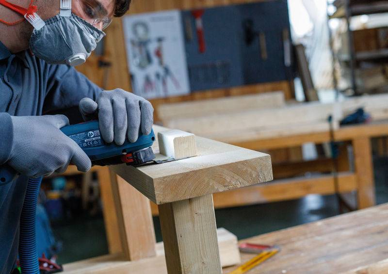 Eine Person, die Schutzausrüstung trägt, verwendet ein Elektrowerkzeug zum Schleifen einer Holzoberfläche in einer Werkstattumgebung.