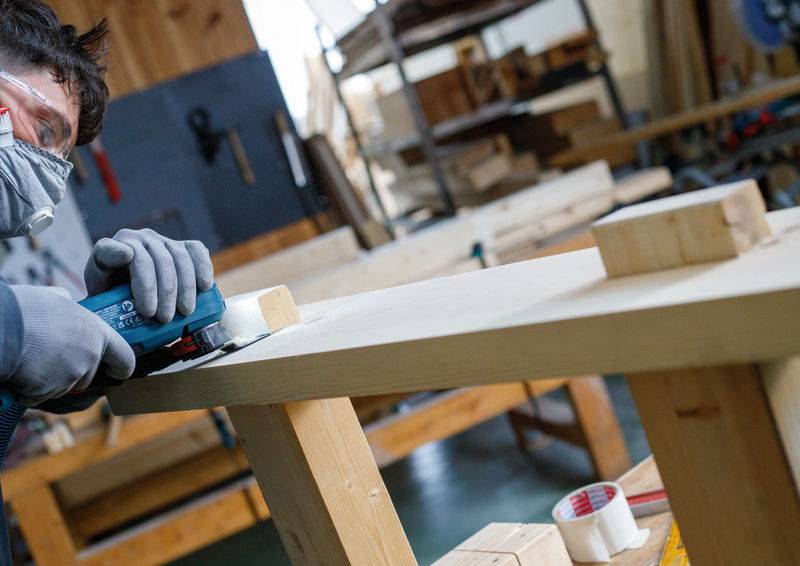 Eine Person in Schutzausrüstung verwendet einen Elektroschleiffer auf einem Holzbrett in einer Werkstatt, die mit Werkzeugen und Werkbänken gefüllt ist.